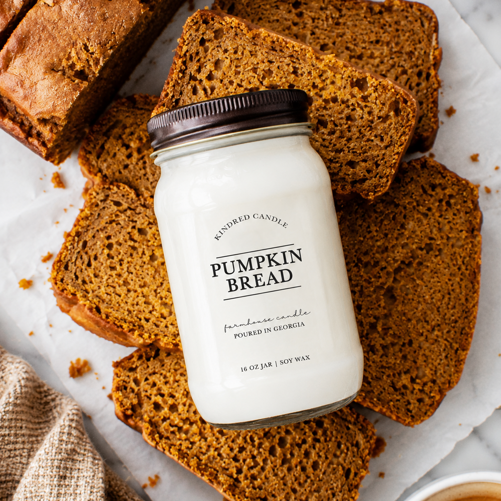 A jar candle labeled 'Pumpkin Bread' by Refined Candle, styled on top of sliced pumpkin bread slices on a white surface with crumbs and a burlap cloth.