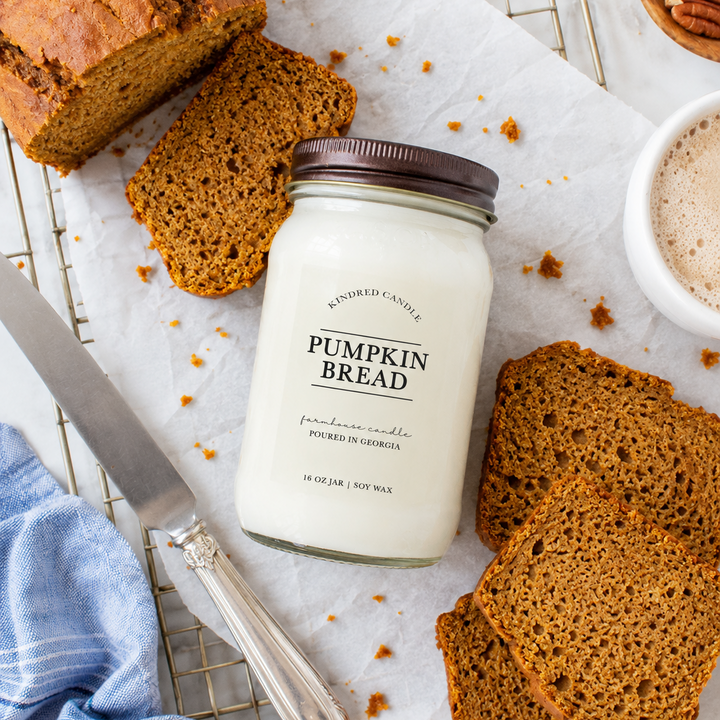 A jar of pumpkin bread scented candle sits on parchment paper surrounded by slices of pumpkin bread, a knife, a blue cloth, and a small bowl of pecans. The candle jar is labeled 'Pumpkin Bread' by 'Whispering Candles'.