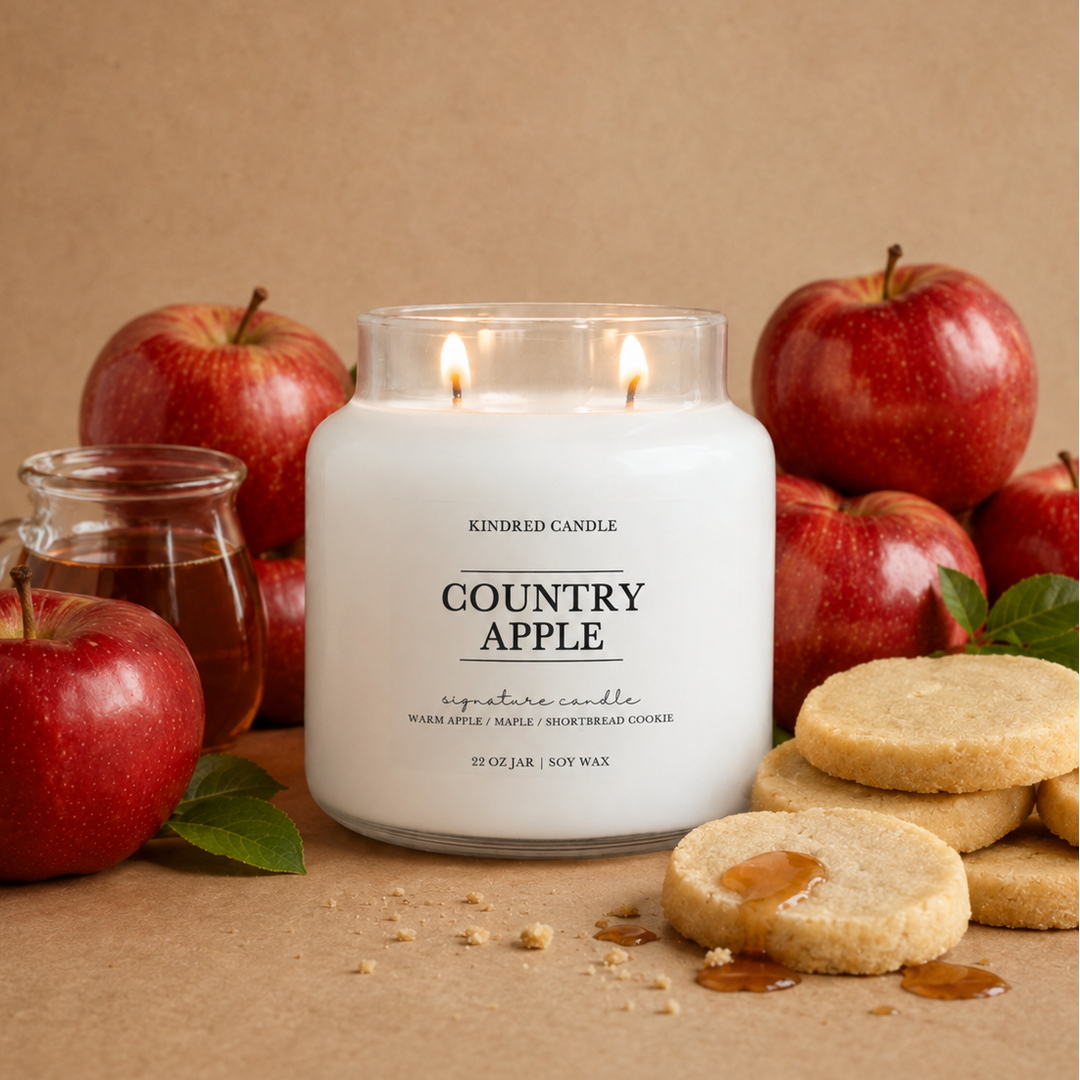 A lit white soy wax candle in a clear glass jar labeled 'COUNTRY APPLE' by Kindred Candle, styled with red apples, sugar cookies, and a small glass jar on a kraft paper background.