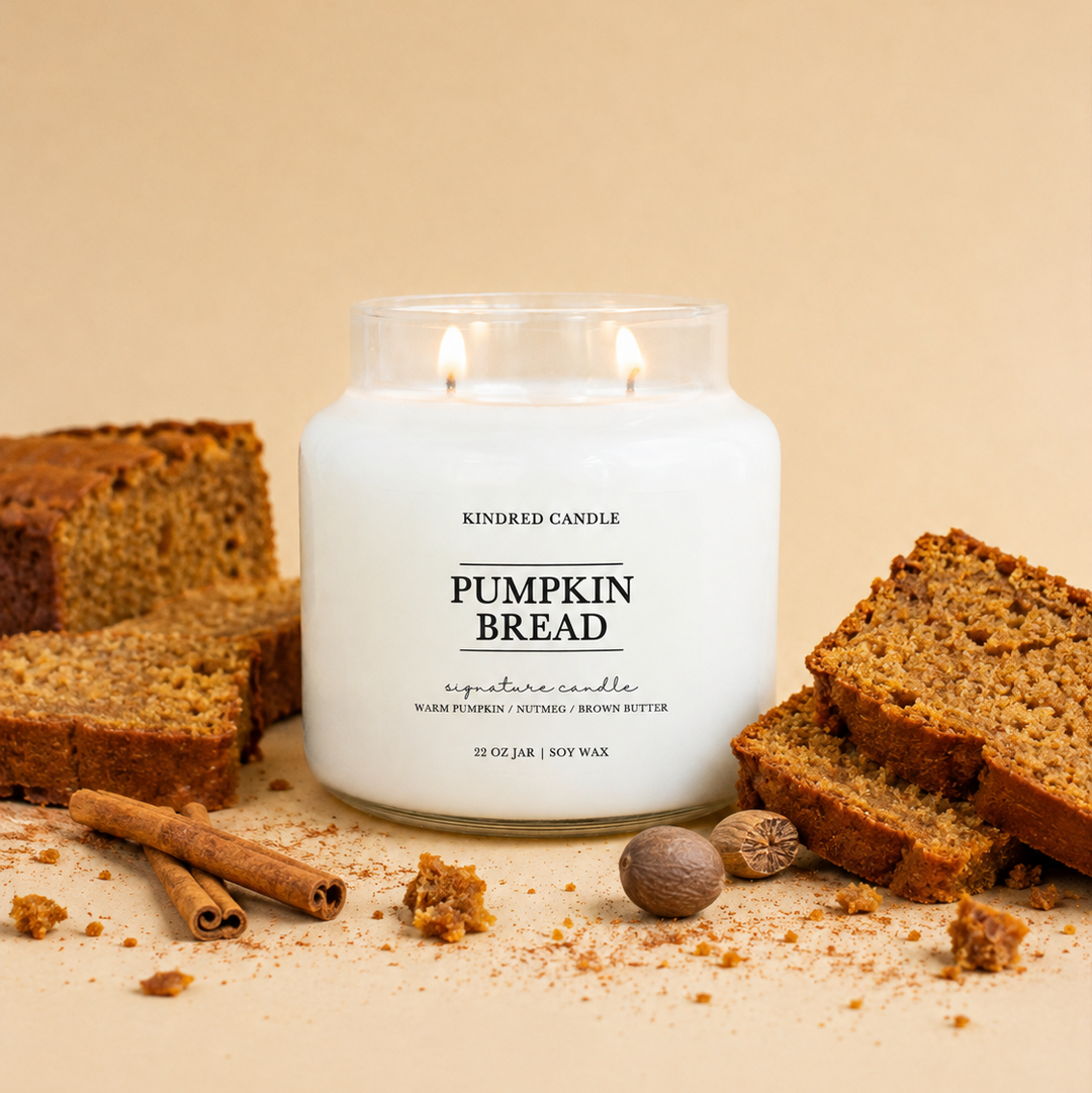 A lit white candle in a clear glass jar labeled 'Pumpkin Bread' by Kindred Candle, styled with sliced pumpkin bread, cinnamon sticks, and nutmeg on a warm beige background.