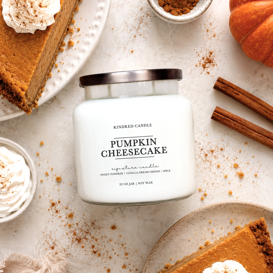 A jar candle labeled 'Pumpkin Cheesecake' by Kindred Candle, styled in a flat lay with pumpkin pie slices, whipped cream, cinnamon sticks, and scattered pumpkin spice on a light neutral background.