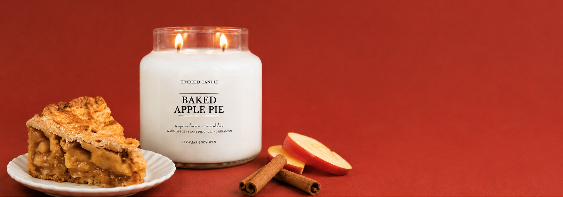 A white jar candle labeled 'BAKED APPLE PIE' by Kindred Candle, styled on a red background with a slice of apple pie on a plate, cinnamon sticks, and apple slices.