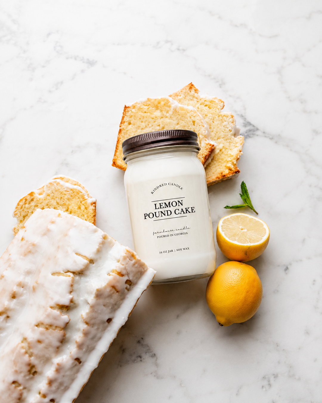 A mason jar candle labeled 'Lemon Pound Cake' by Kindred Candle styled on a white marble surface with lemon slices, a whole lemon, and pieces of lemon pound cake around it.