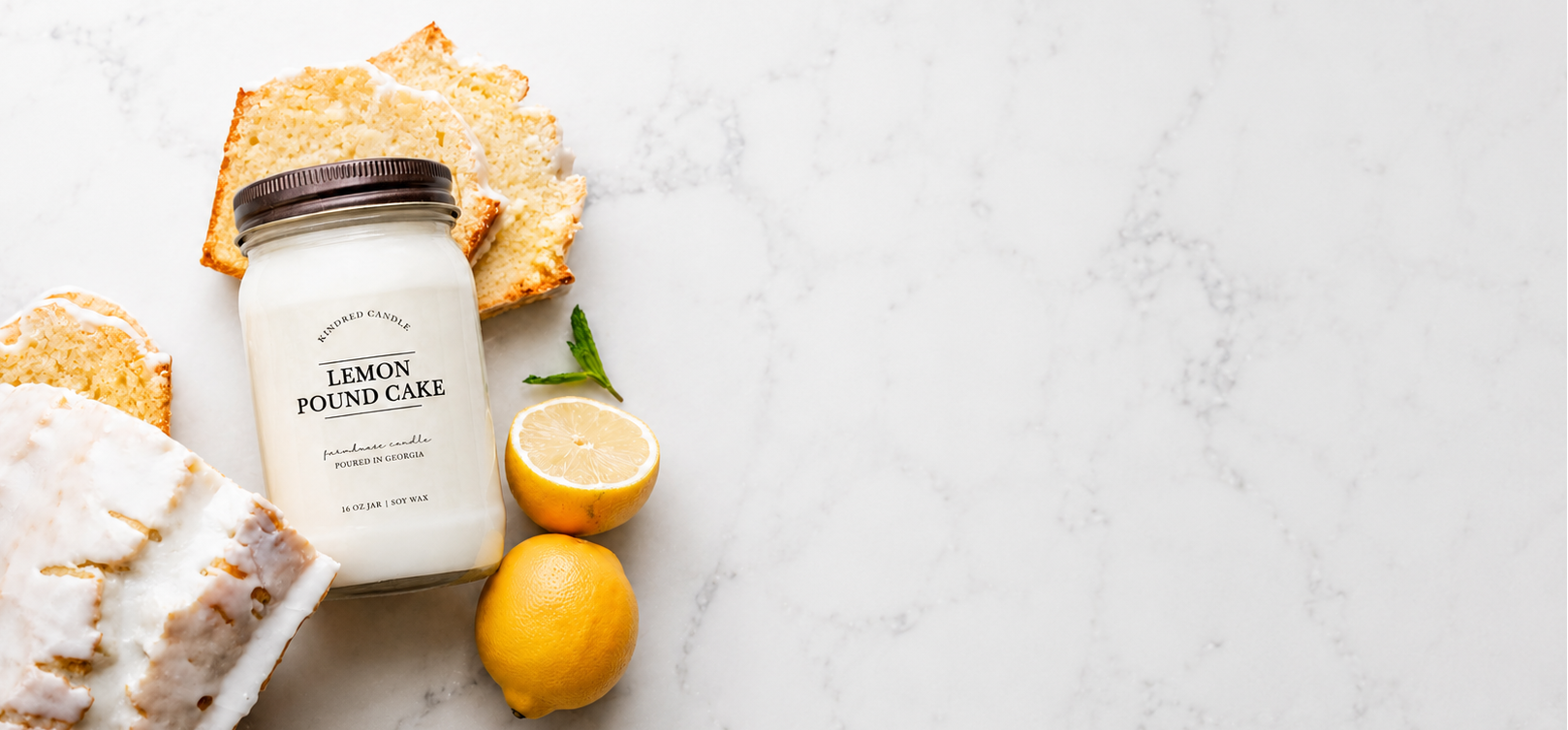 A Kindred Candle jar candle labeled 'LEMON POUND CAKE' sits on a light marble surface with lemon slices, a piece of cake, and graham crackers. The candle is in a clear glass jar with white wax and a dark lid.