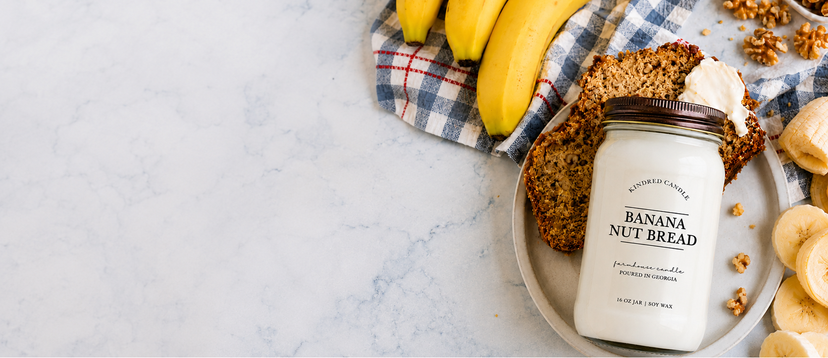 a mason jar soy candle labeled Banana Nut Bead from Kindred Candle lays flat on a marble counter with slices of banana bread, walnuts, and bananas from Kindred Candle