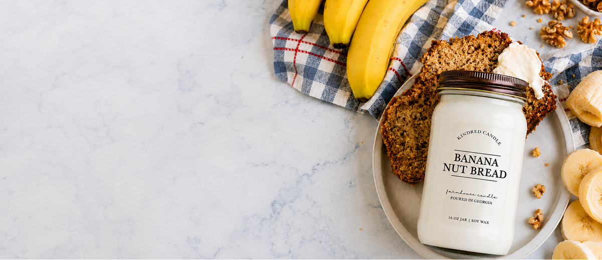 a mason jar soy candle labeled Banana Nut Bead from Kindred Candle lays flat on a marble counter with slices of banana bread, walnuts, and bananas from Kindred Candle