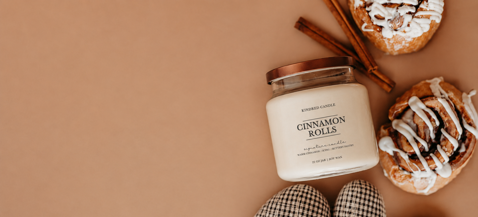 A jar candle labeled 'CINNAMON ROLLS' by Kindred Candle, styled in a flat lay with cinnamon rolls, cinnamon sticks, and plaid oven mits on a warm tan background.