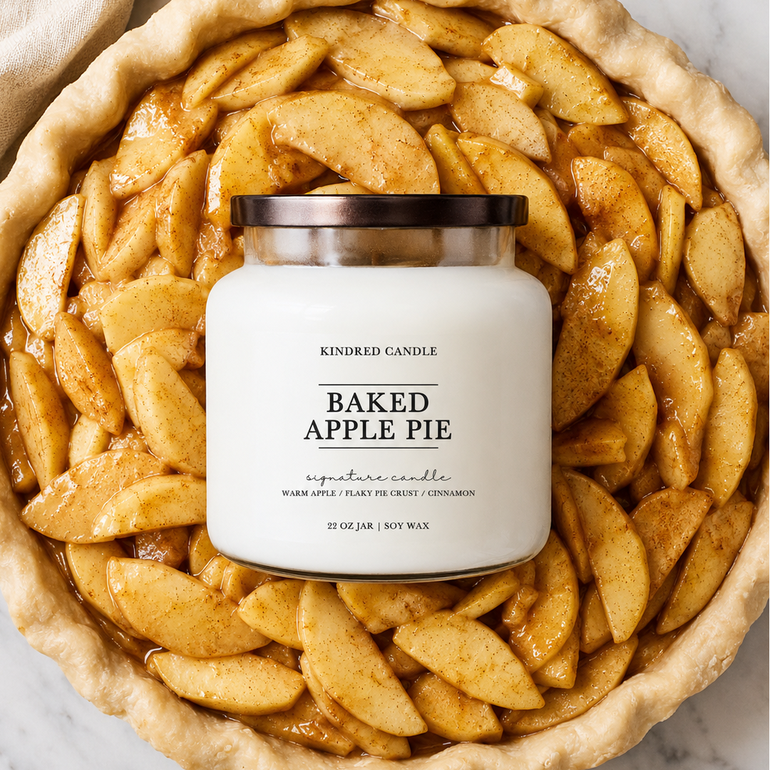 A jar candle labeled 'BAKED APPLE PIE' by Kindred Candle, placed on top of a baked apple pie crust with sliced apples, photographed from above on a light marble surface.