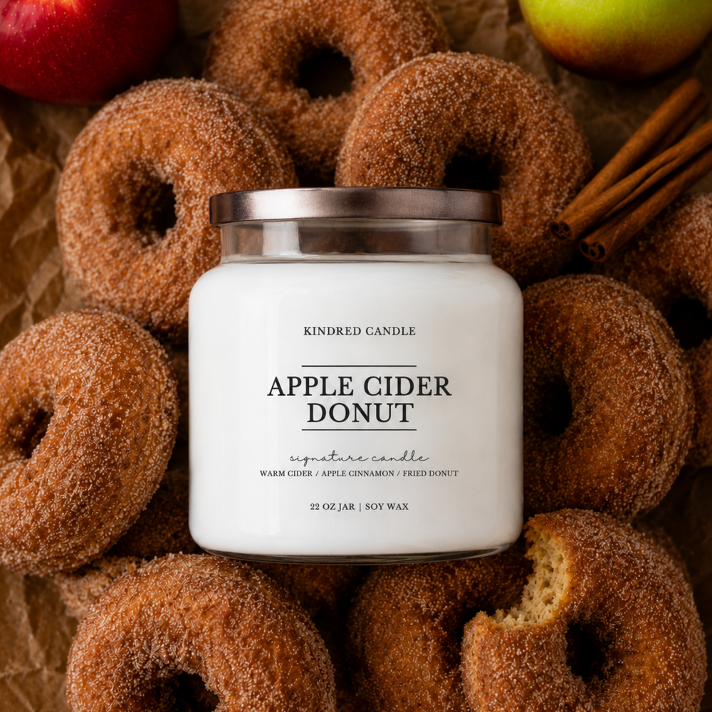 A jar candle labeled 'Apple Cider Donut' by Kindred Candle, styled on brown paper with sugar-coated donuts, cinnamon sticks, and red and green apples around it.