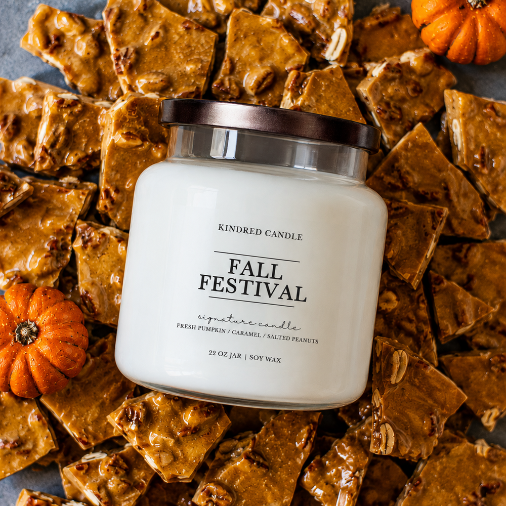 A white jar candle labeled 'KINDRED CANDLE FALL FESTIVAL' sits on top of autumn-themed candy bark pieces with small orange pumpkins around it. The candle is shown in a styled flat-lay arrangement.