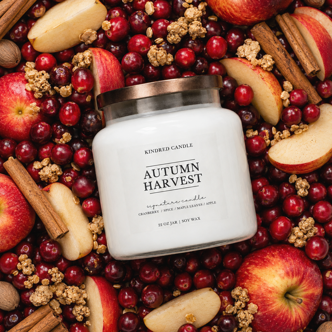 A white jar candle labeled 'Autumn Harvest' by Kindred Candle, surrounded by red apples, cranberries, cinnamon sticks, and granola in a flat-lay arrangement.