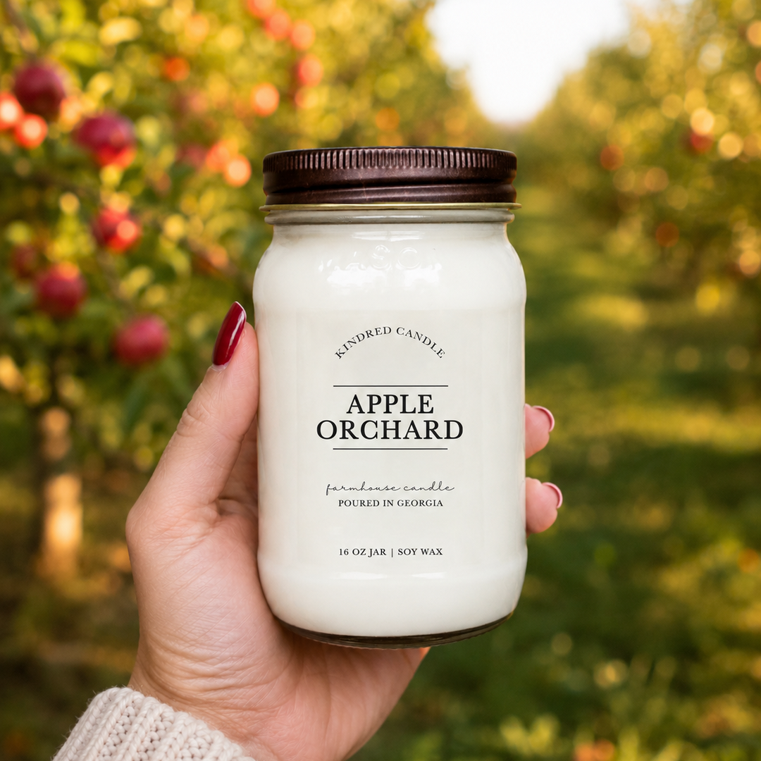 A hand holds a 16 oz mason jar candle labeled 'Apple Orchard' by Kindred Candle against a blurred outdoor background of an apple orchard.