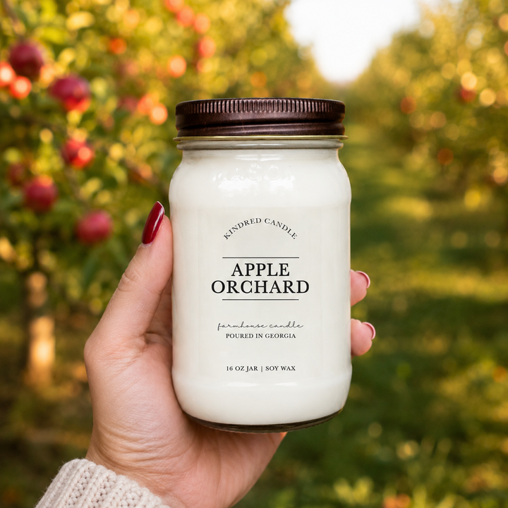 A hand holds a 16 oz mason jar candle labeled 'Apple Orchard' by Kindred Candle against a blurred outdoor background of an apple orchard.