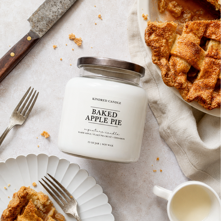 A white glass jar candle labeled 'Baked Apple Pie' by Kindred Candle, styled in a flat lay with a sliced apple pie, a knife, forks, a plate, and a small cup on a light neutral surface.