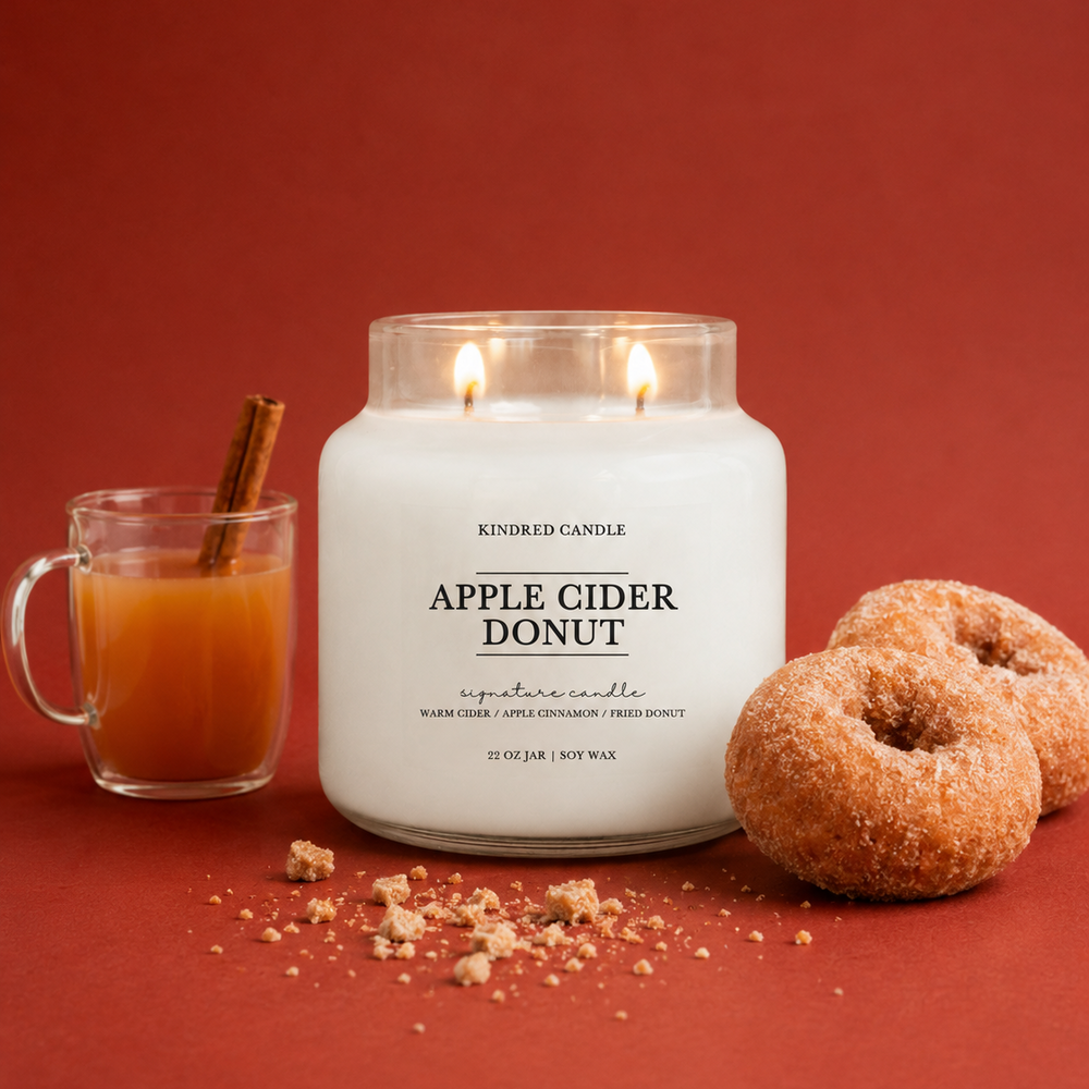 A lit white candle in a clear glass jar labeled 'APPLE CIDER DONUT' by Kindred Candle, styled on a warm red background with a glass mug of orange drink and cinnamon stick, plus two sugar-topped donuts.