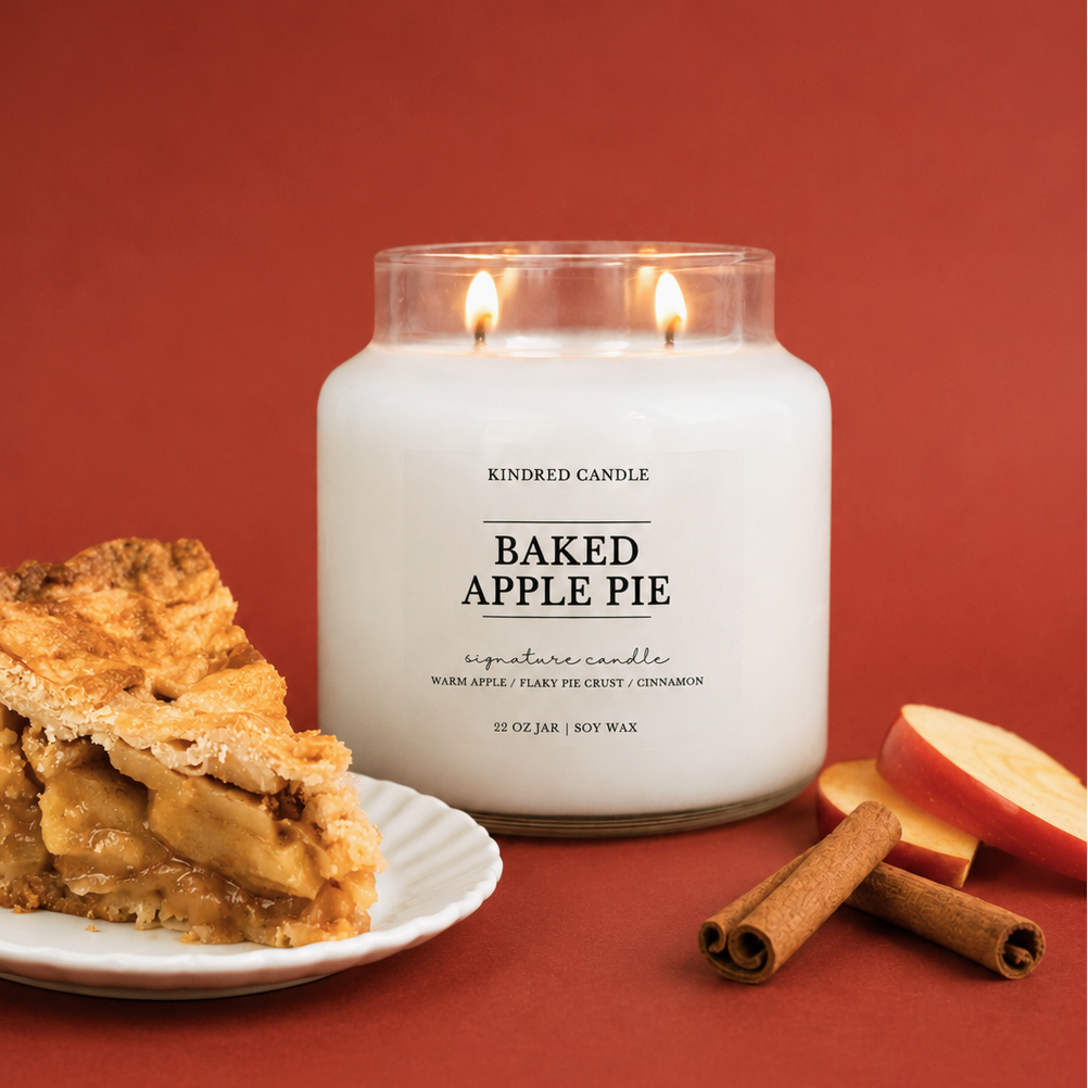 A lit white candle in a clear glass jar labeled 'KINDRED CANDLE BAKED APPLE PIE' sits on a red background with a slice of apple pie on a plate, cinnamon sticks, and apple slices nearby.