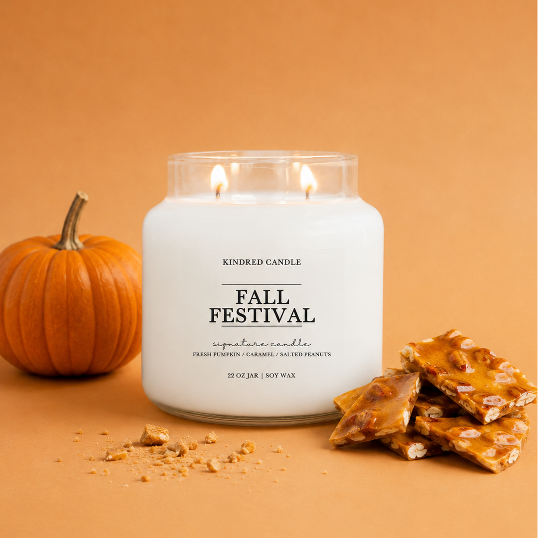 A white jar candle labeled 'FALL FESTIVAL' by Kindred Candle, styled on a warm orange-brown background with a small pumpkin on the left and caramel-like decorative pieces on the right.