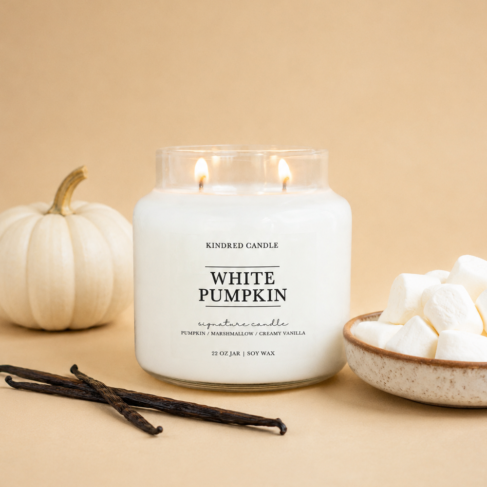 A lit white soy wax candle in a clear glass jar labeled 'White Pumpkin' by Kindred Candle, styled on a beige background with a small white pumpkin, vanilla beans, and marshmallows in a bowl.