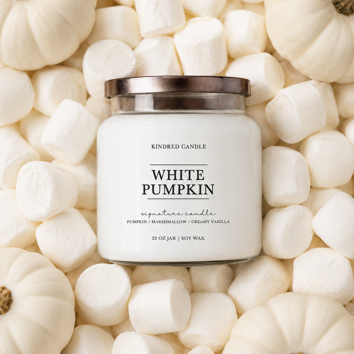 A white scented candle in a clear glass jar with a metallic lid, surrounded by white pumpkins and marshmallows. The label reads 'KINDRED CANDLE' and 'WHITE PUMPKIN'.