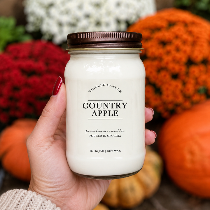 A hand with red nail polish holds a 16 oz jar candle labeled 'COUNTRY APPLE' by Kindred Candle. The candle is in a clear glass jar with white wax and a brown metal lid, set against a blurred autumn background of pumpkins and flowers.