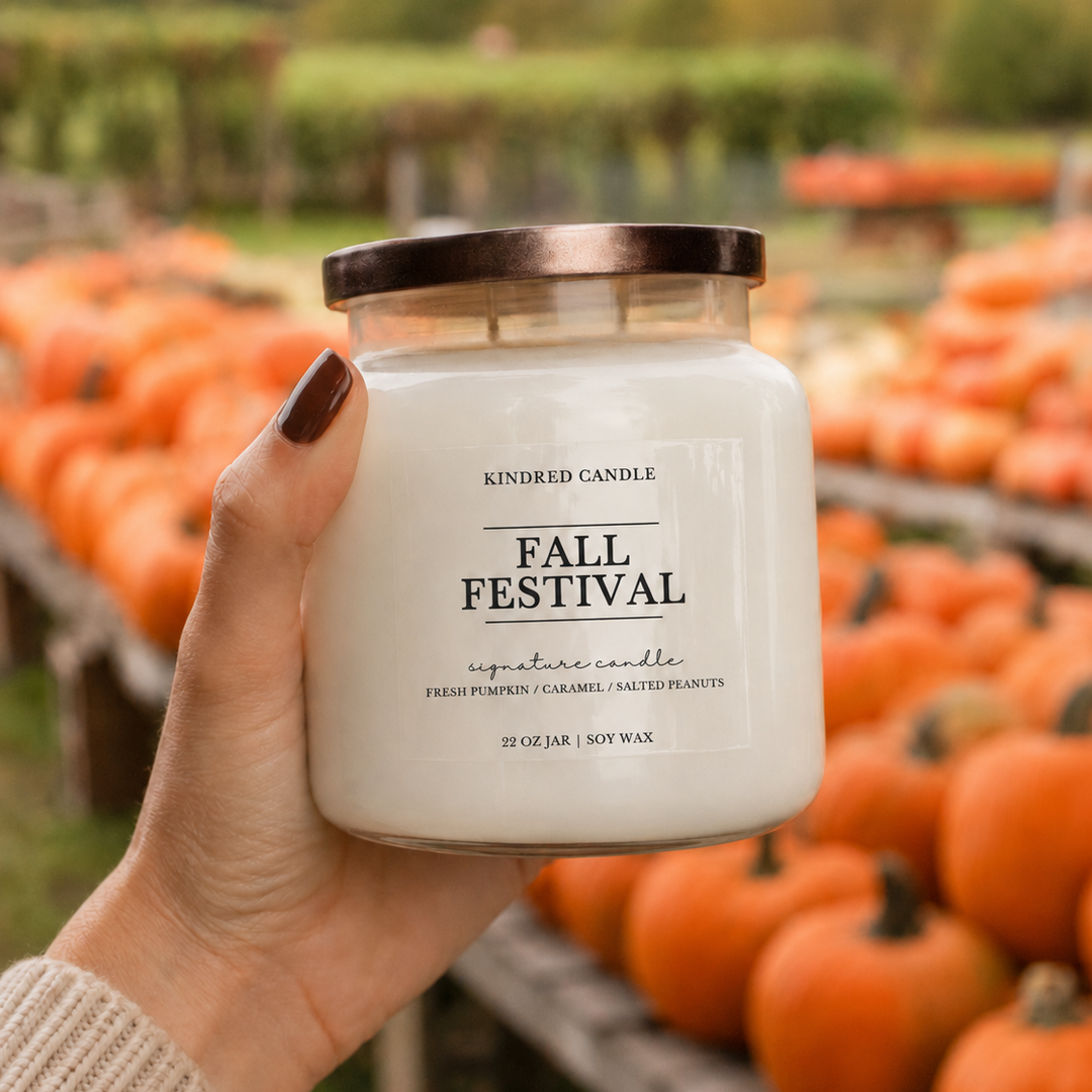 A hand holds a jar candle labeled 'KINDRED CANDLE FALL FESTIVAL' in front of a blurred outdoor pumpkin patch with rows of orange pumpkins.