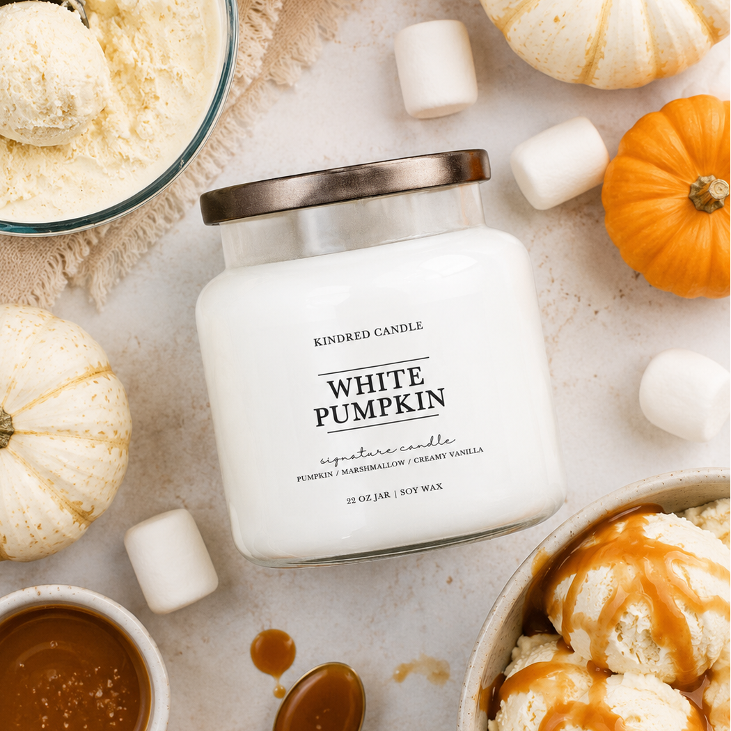 A white candle jar labeled 'White Pumpkin' by Kindred Candle, styled in a fall-themed flat lay with small pumpkins, ice cream scoops, marshmallows, a bowl of caramel sauce, and a spoon on a light neutral surface.