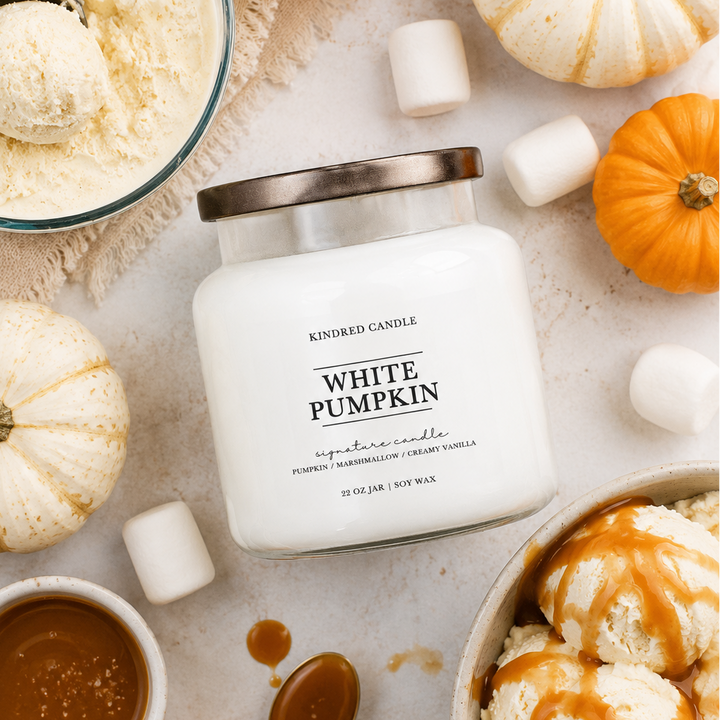 A white candle jar labeled 'White Pumpkin' by Kindred Candle, styled in a fall-themed flat lay with small pumpkins, ice cream scoops, marshmallows, a bowl of caramel sauce, and a spoon on a light neutral surface.