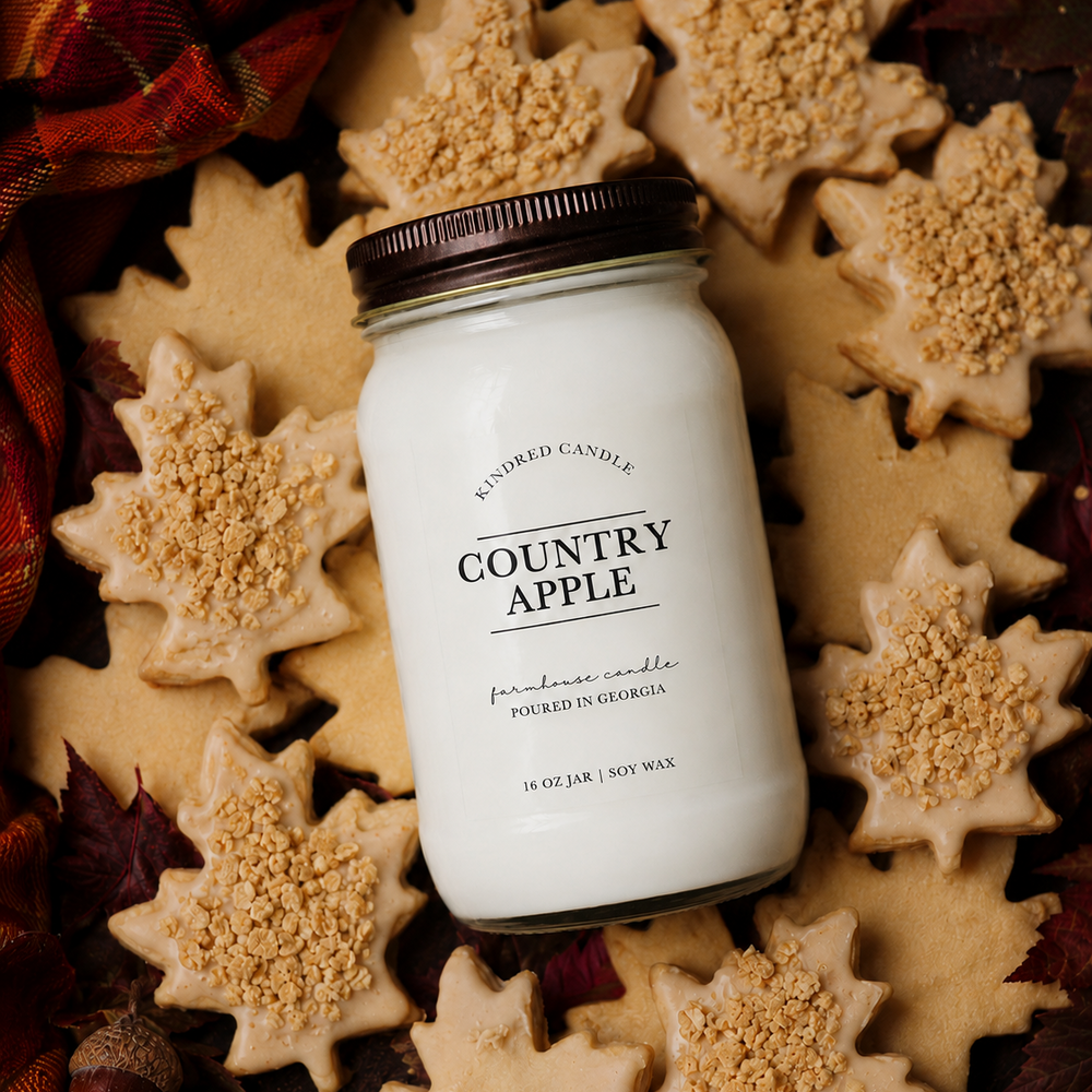 A 16 oz glass jar candle labeled 'COUNTRY APPLE' by Kindred Candle, styled in a flat lay with star-shaped sugar cookies and autumn leaves around it.