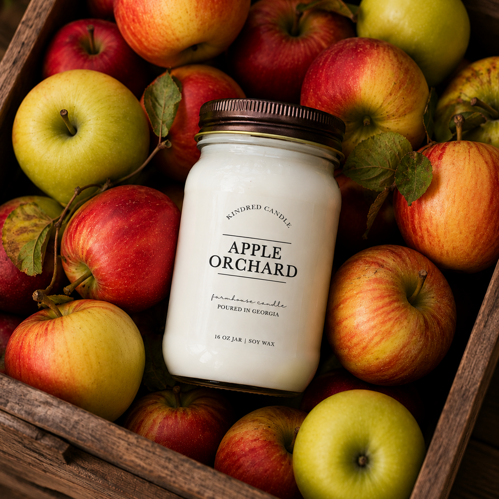 A jar candle labeled 'Apple Orchard' by Kindred Candle, placed in a rustic wooden crate filled with red and green apples.