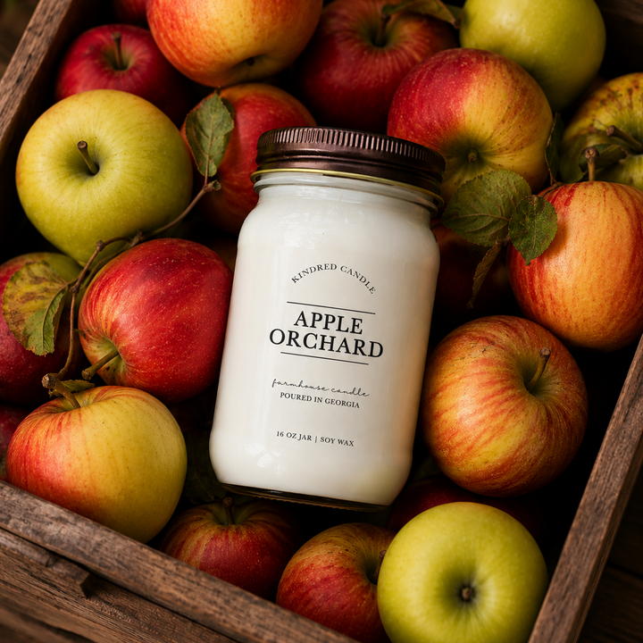 A jar candle labeled 'Apple Orchard' by Kindred Candle, placed in a rustic wooden crate filled with red and green apples.