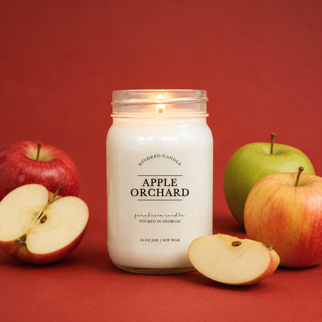 A lit white candle in a clear glass mason jar labeled 'APPLE ORCHARD' by Kindred Candle, styled with whole and sliced red and green apples on a solid dark red background.