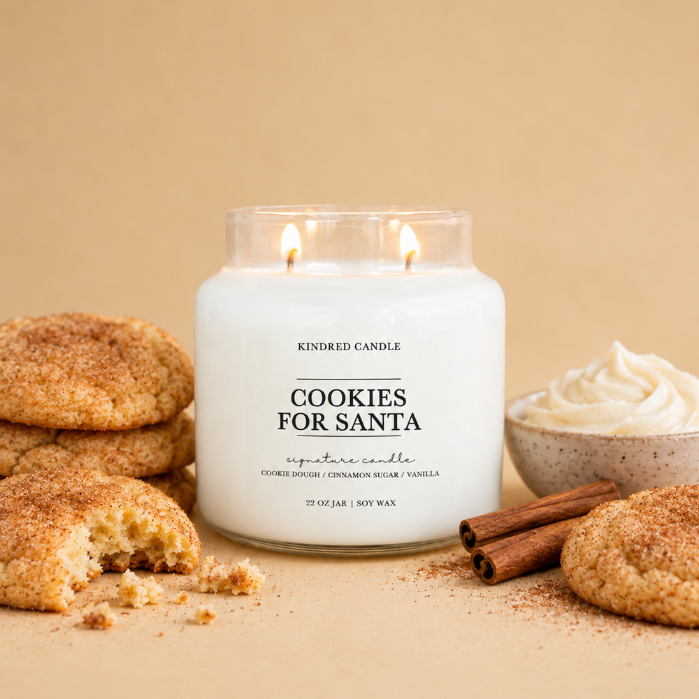 A lit soy candle in a clear glass jar labeled 'Cookies for Santa' by Kindred Candle, styled with snickerdoodle cookies, cinnamon sticks, and whipped cream on a warm beige background.