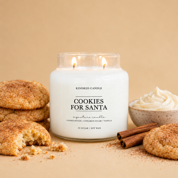 A lit soy candle in a clear glass jar labeled 'Cookies for Santa' by Kindred Candle, styled with snickerdoodle cookies, cinnamon sticks, and whipped cream on a warm beige background.