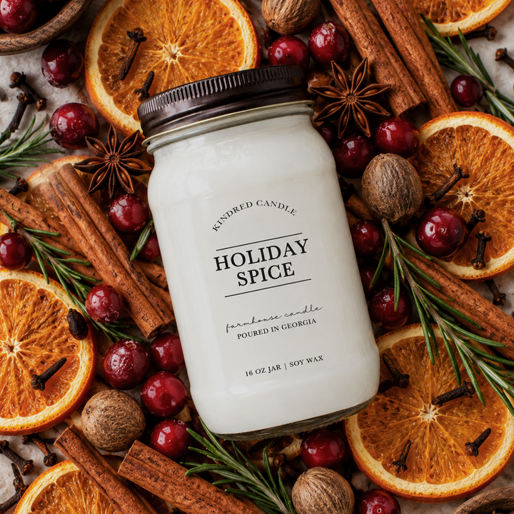 A mason jar, soy candle labeled 'Holiday Spice' by Kindred Candle, styled in a flat lay with cinnamon sticks, dried orange slices, cranberries, star anise, and clove.