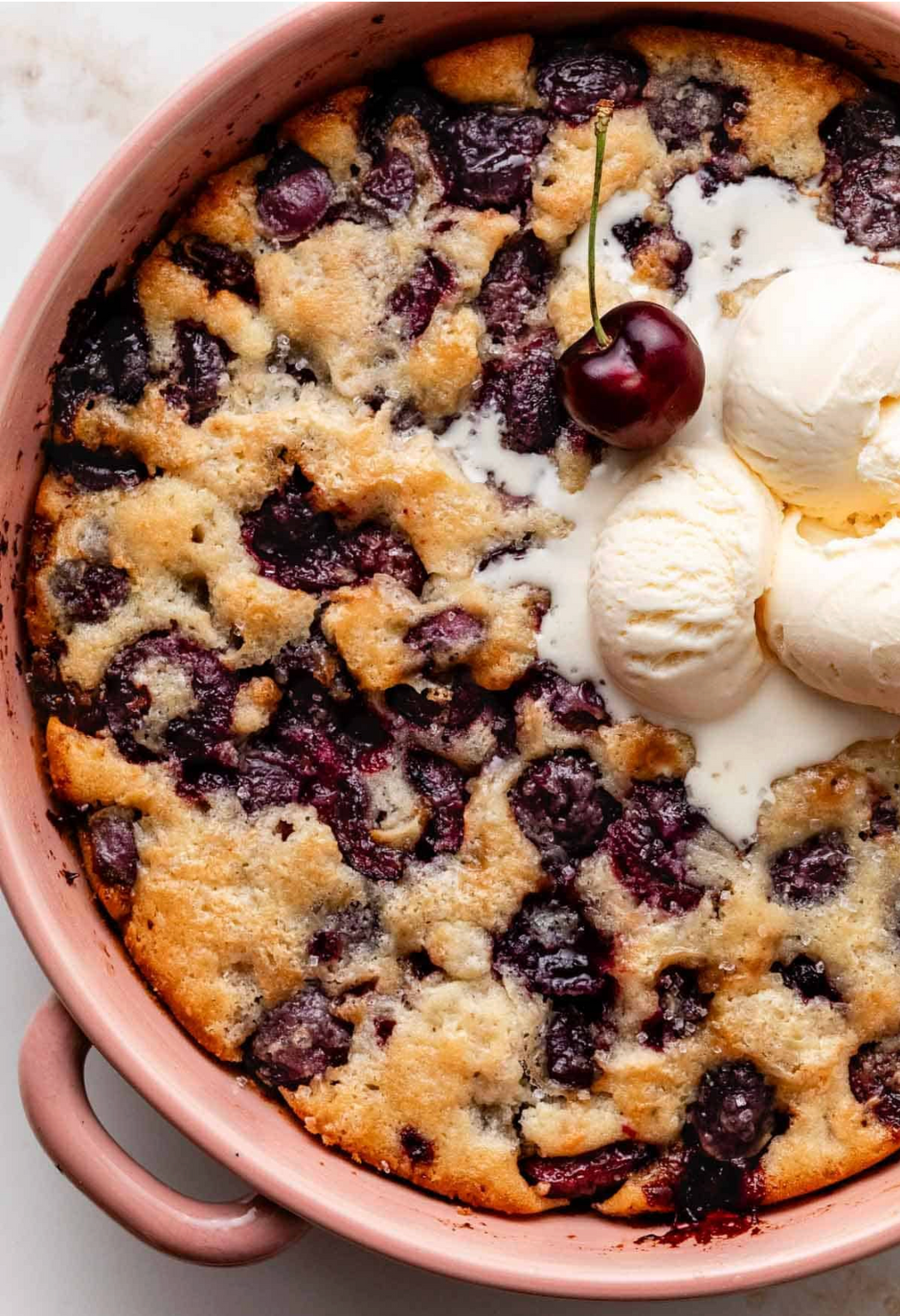 A close-up overhead shot of a baked berry cobbler in a pink ceramic dish, topped with vanilla ice cream and a single fresh cherry on a light neutral background.