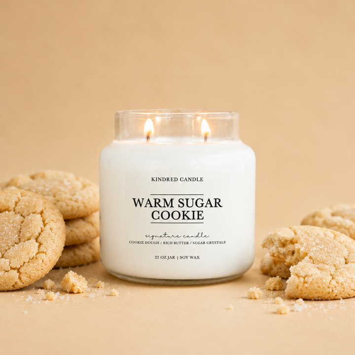 A soy scented candle in a clear glass jar labeled 'Warm Sugar Cookie' by Kindred Candle, styled with sugar-coated cookies on a warm beige background.